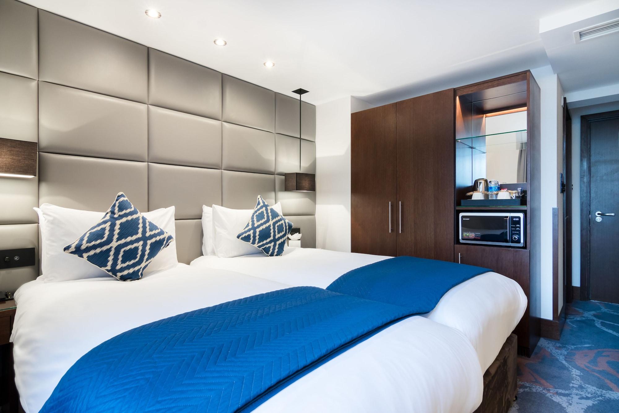 Montcalm Chilworth Townhouse, Paddington Hotel London Ngoại thất bức ảnh The photo shows a modern hotel room featuring two beds with white linens and decorative blue throw blankets. Each bed has a pair of patterned cushions. The walls behind the beds are adorned with a sleek, padded design. To the side, there’s a wooden c