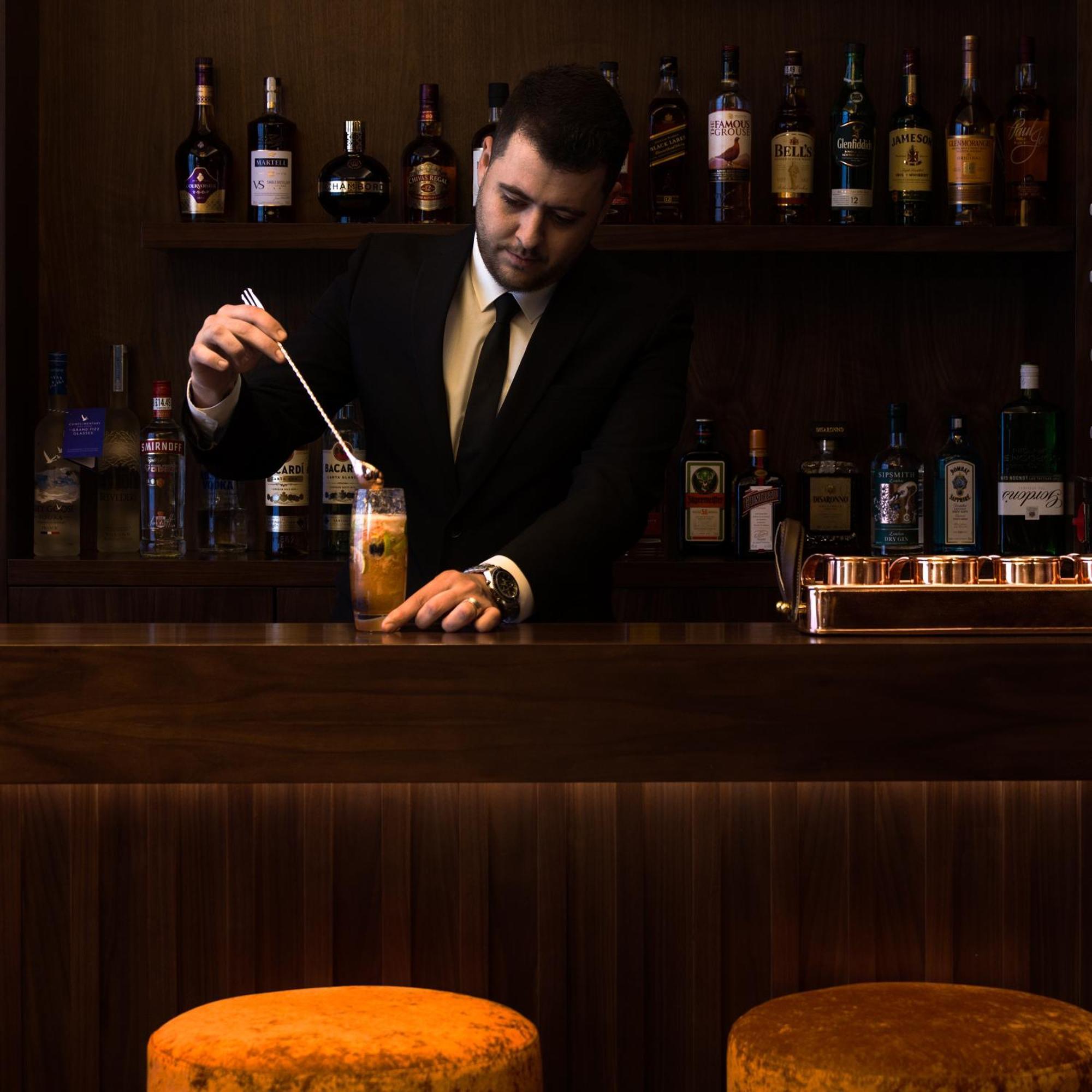 Montcalm Chilworth Townhouse, Paddington Hotel London Ngoại thất bức ảnh The image shows a bartender at a bar. He is wearing a black suit and tie and is focused on preparing a cocktail, stirring it with a spoon. Behind him, there is a wooden bar counter and a shelf filled with various bottles of liquor and spirits. The at